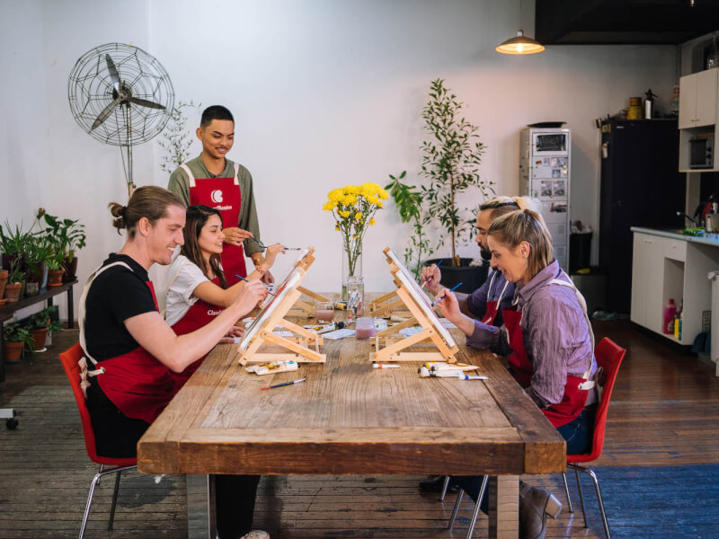A group of men and women smile as they paint on canvases around a table